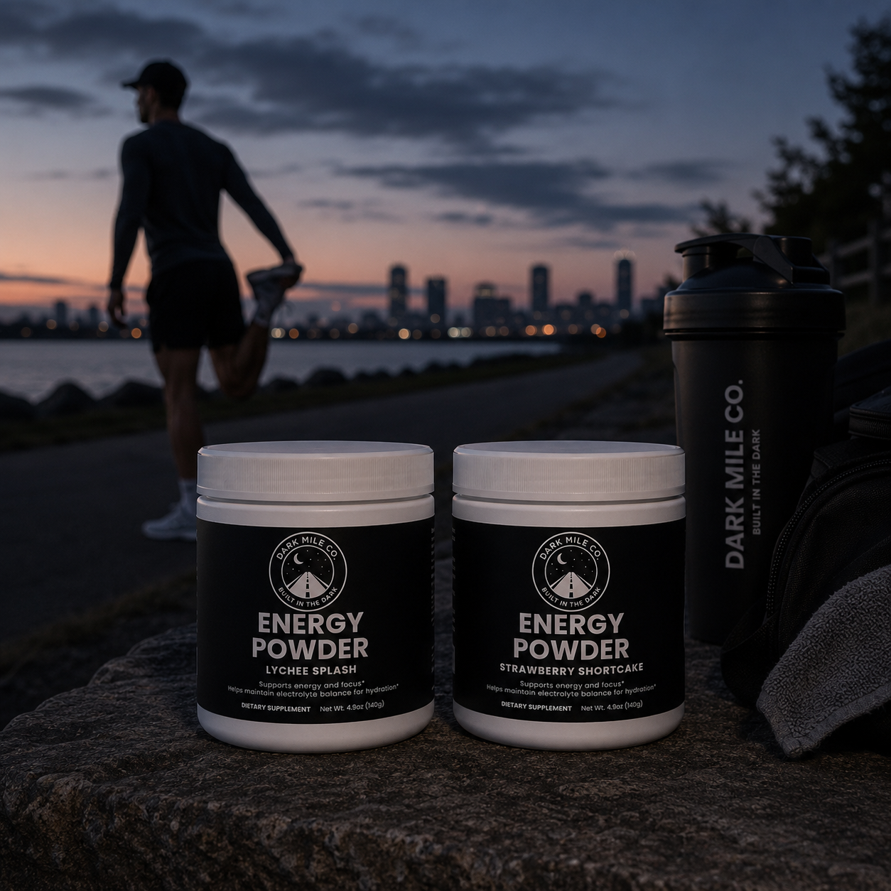 Dark Mile Co. Energy Powder supplements in Lychee Splash and Strawberry Shortcake flavors displayed on a stone ledge before sunrise, with a runner stretching along a waterfront path, a shaker bottle, and a city skyline in the background.