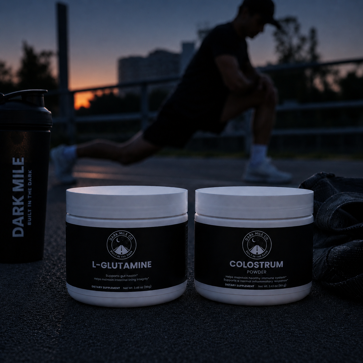 Dark Mile Co. L-Glutamine and Colostrum powders on pavement at pre-dawn, with a shaker bottle, gym bag, and runner stretching in the dark background.